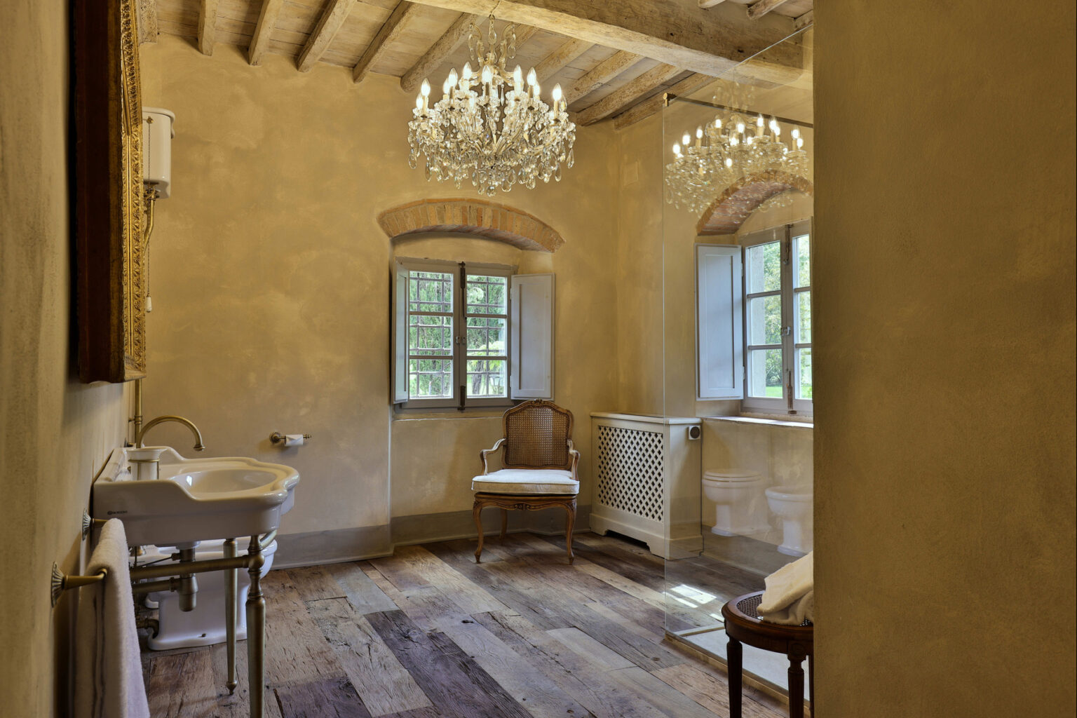 Elegant rustic bathroom featuring wooden flooring, vintage washbasin, and two ornate chandeliers hanging from the ceiling, complemented by natural light from large windows. | Only on SopranoVillas