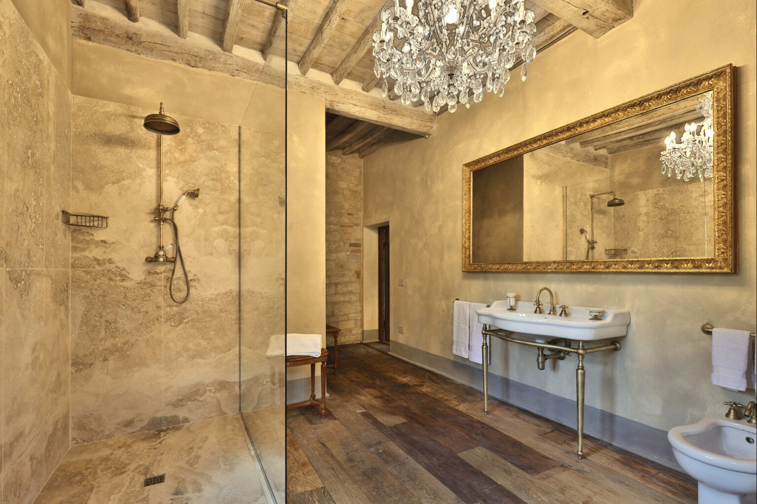 Elegant modern bathroom featuring a glass shower, vintage chandelier, and a large ornate mirror over a stylish sink. The warm wooden flooring complements the textured beige walls, creating a luxurious atmosphere. | Only on SopranoVillas