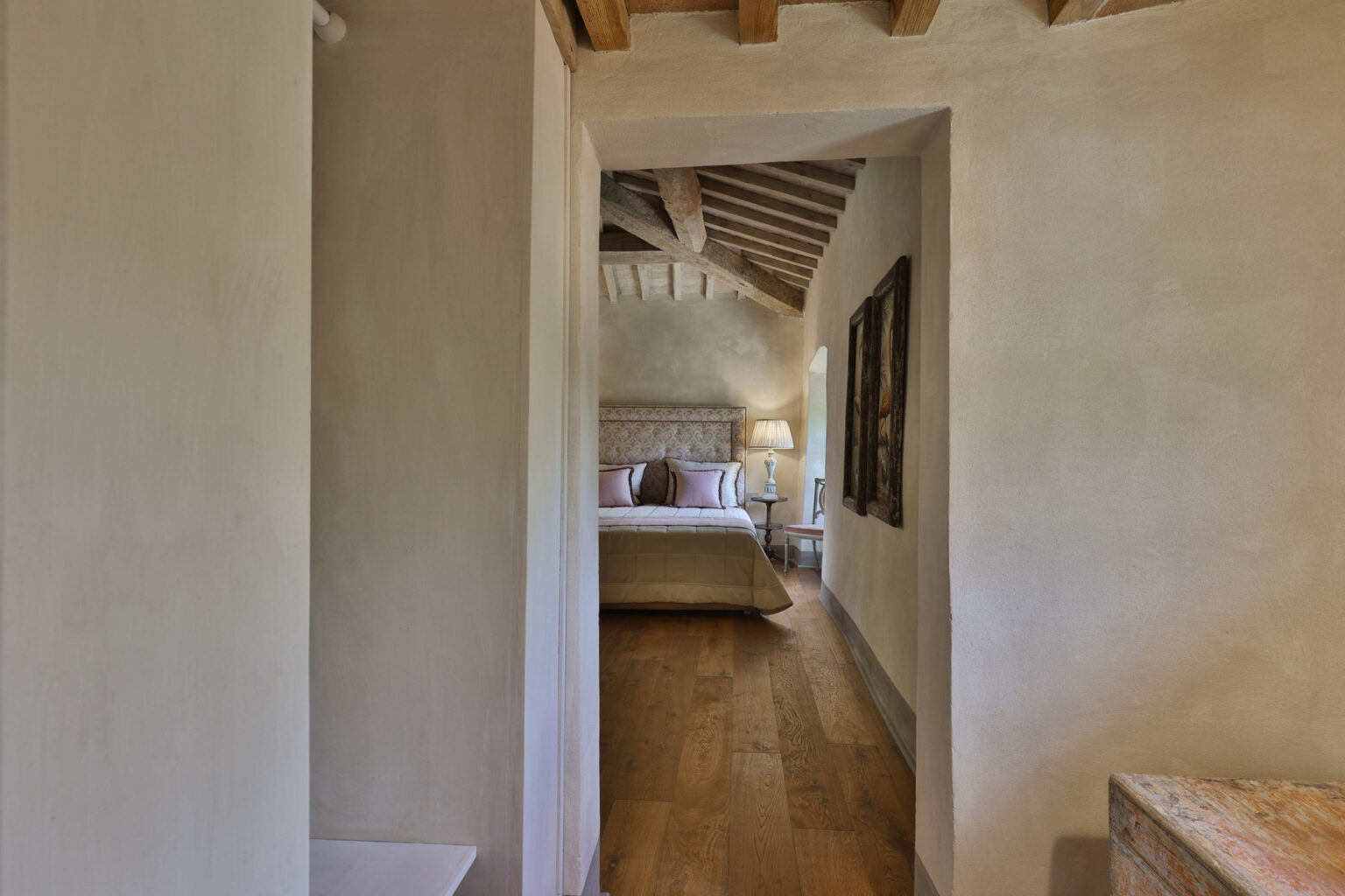 Interior view of a cozy bedroom with a wooden ceiling, featuring a king-sized bed, soft lighting, and decorative artwork, framed by a hallway with a neutral color palette and hardwood flooring. | Only on SopranoVillas