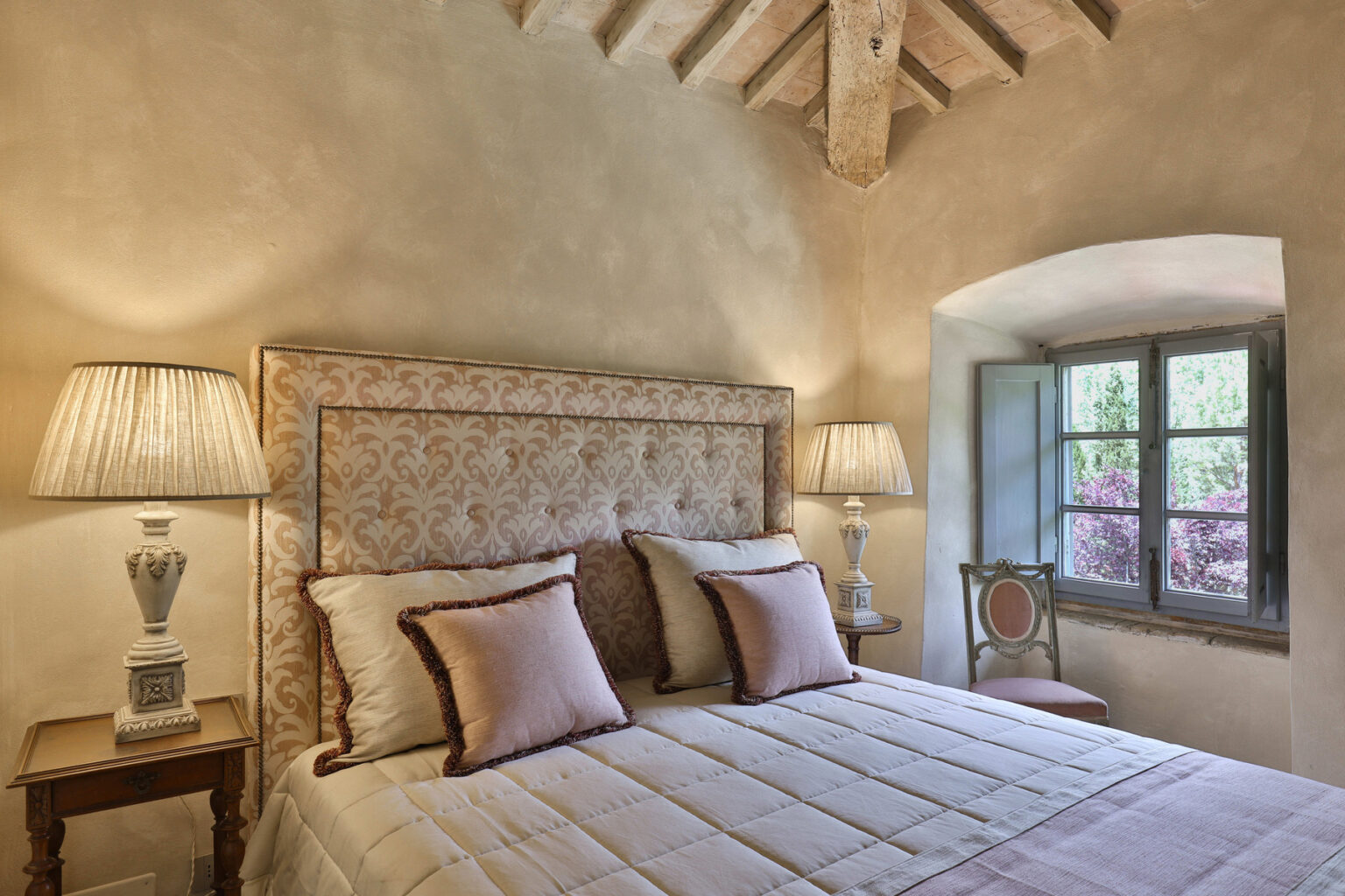 Cozy bedroom interior featuring a plush bed with decorative pillows, elegant bedside lamps, and a charming window that offers natural light and views of greenery. The rustic style is enhanced by wooden beams and soft color tones, creating a serene atmosphere. | Only on SopranoVillas