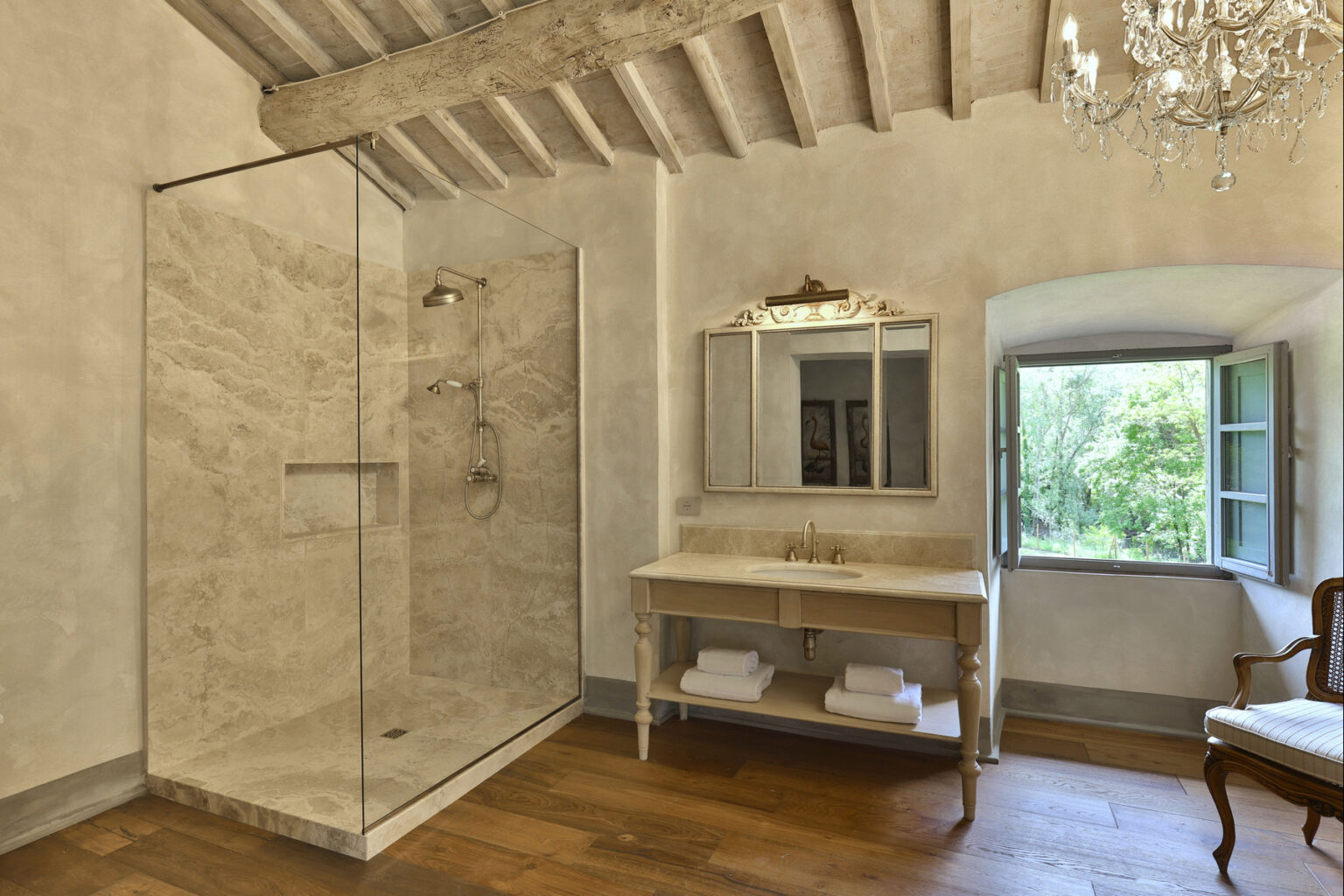Modern bathroom featuring a spacious glass shower, elegant wooden flooring, and a vintage vanity with a large mirror. Natural light streams in through an open window, enhancing the serene atmosphere. | Only on SopranoVillas
