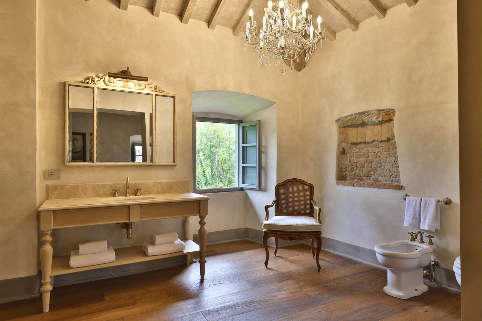 Elegant bathroom interior featuring a vintage-style wooden vanity with a marble sink, a large ornate mirror, and a stylish chandelier. Natural light streams through a window, illuminating the warm wooden floor and a classic chair. Decorative elements include neatly stacked towels and a charming exposed brick wall. | Only on SopranoVillas
