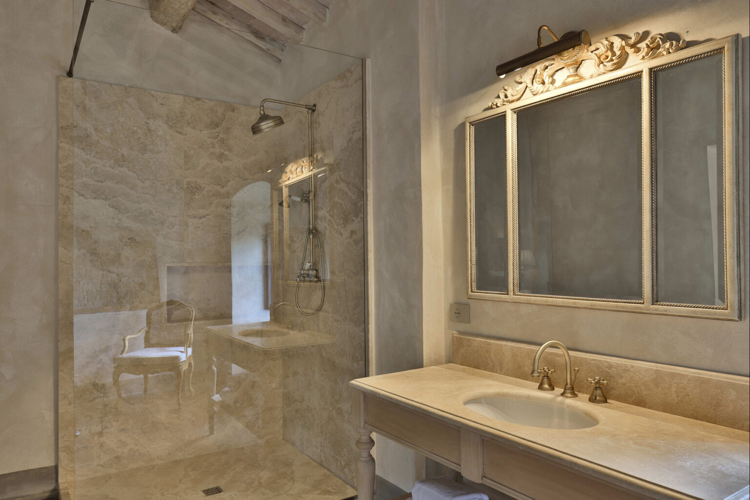 Luxurious bathroom featuring a marble walk-in shower with a rainfall showerhead, a wooden vanity with a sink and elegant gold fixtures, and a decorative mirror illuminated by a stylish light fixture. The neutral color palette and natural textures create a serene atmosphere. | Only on SopranoVillas