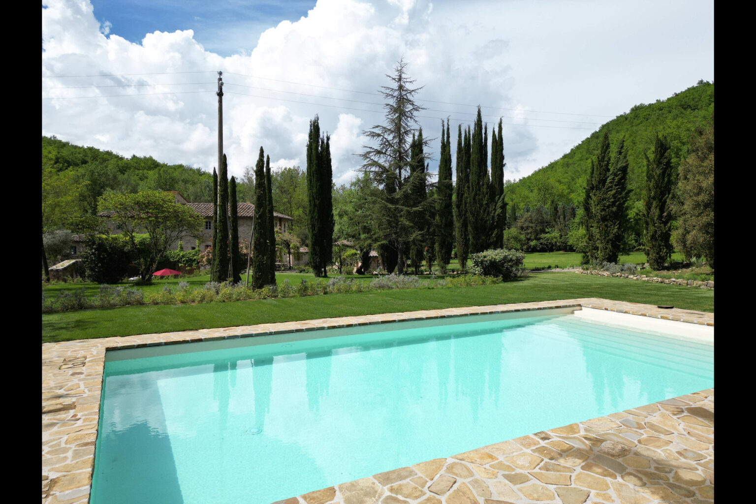 Scenic view of a clear swimming pool surrounded by lush greenery, tall cypress trees, and a charming stone villa under a blue sky with fluffy clouds. Ideal for relaxation and outdoor living. | Only on SopranoVillas