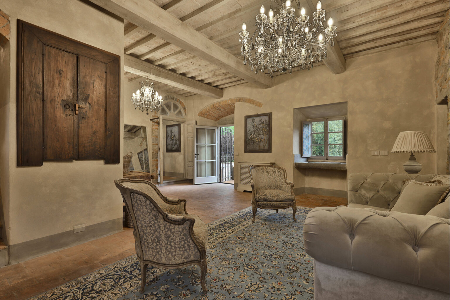 Elegant living room featuring a vintage design with exposed wooden beams, two ornate armchairs, and a plush tufted sofa. The room is illuminated by two crystal chandeliers, enhancing the sophisticated ambiance. Natural light streams through the window and French doors, which open to an outdoor view. A decorative area rug adds warmth to the terracotta floor, while artwork adorns the walls, contributing to the room's charm. | Only on SopranoVillas