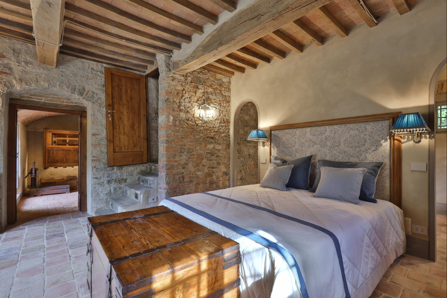 Cozy bedroom featuring a rustic design with exposed brick walls, wooden beams, and a comfortable bed adorned with decorative pillows. The room includes a vintage wooden trunk at the foot of the bed and stylish blue lamps, creating a warm and inviting atmosphere. A doorway leads to an adjacent area with stairs, enhancing the room's charm. | Only on SopranoVillas