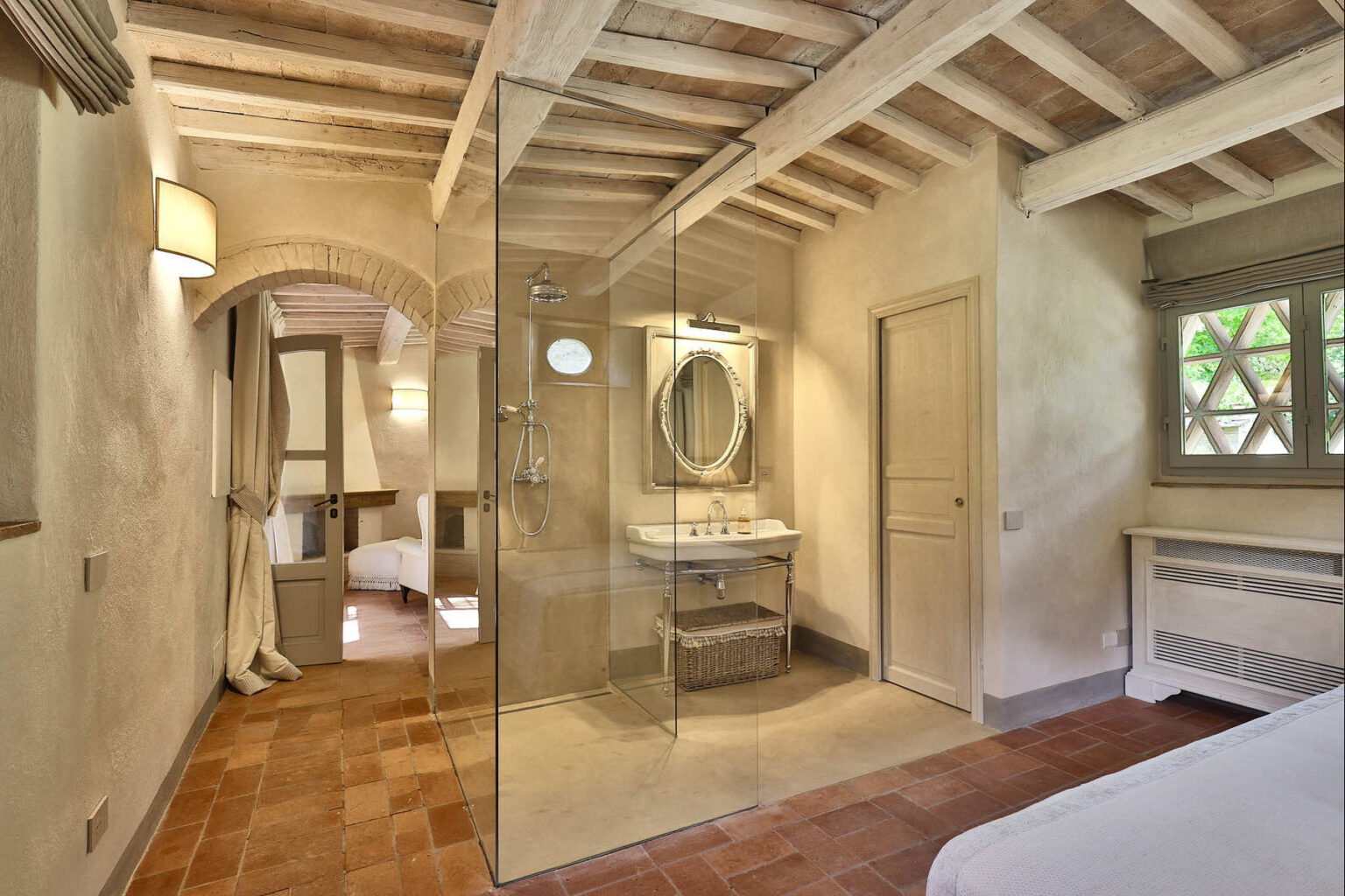 Modern bedroom with an en-suite bathroom featuring a glass shower, wooden ceiling beams, and warm-toned tile flooring, creating a cozy and inviting atmosphere. | Only on SopranoVillas
