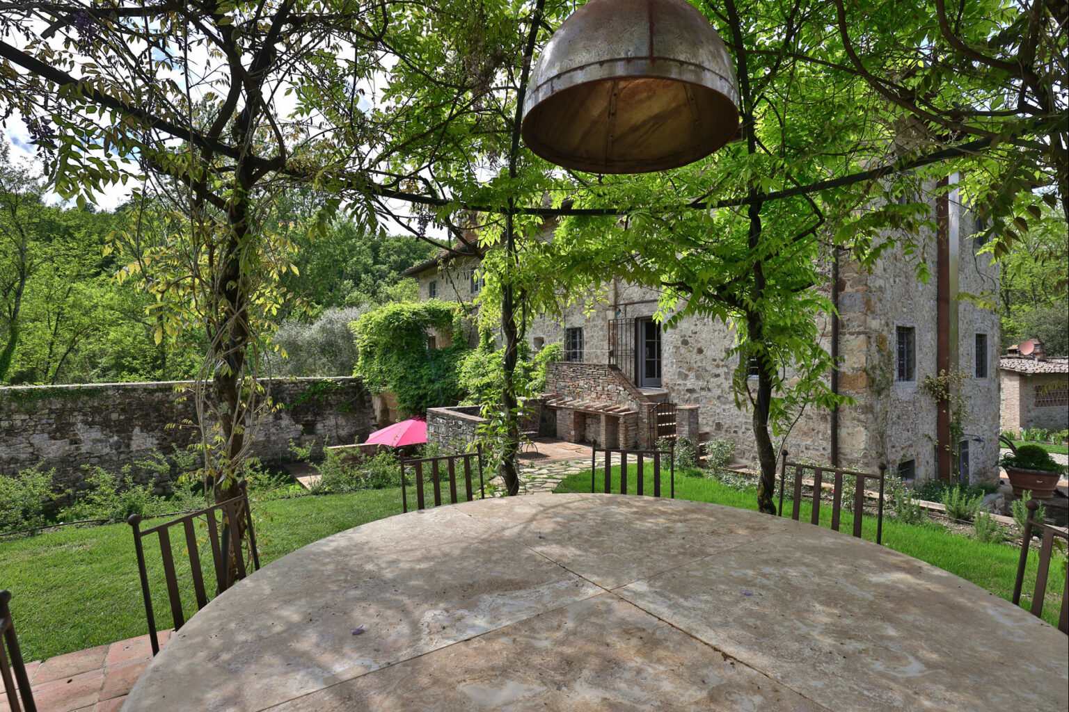 Rustic outdoor dining area with a round table surrounded by chairs, framed by lush greenery and a stone building in the background, creating a serene countryside atmosphere. | Only on SopranoVillas
