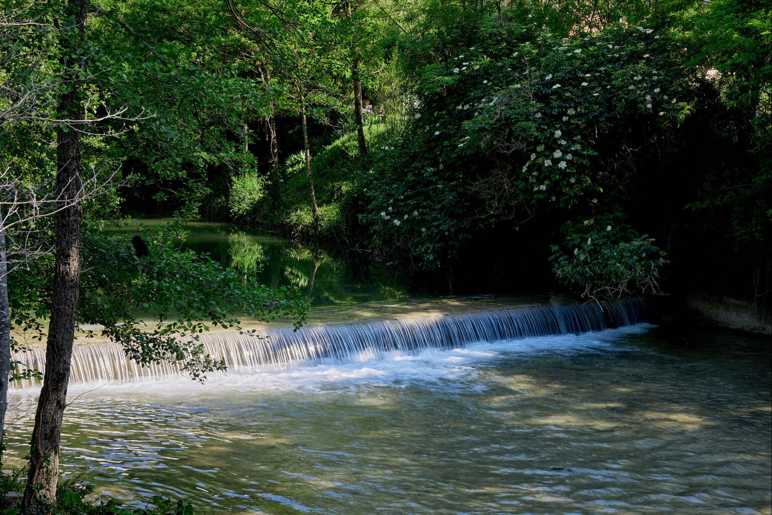 Tranquil waterfall cascading over rocks into a serene river, surrounded by lush greenery and trees, creating a peaceful natural landscape. | Only on SopranoVillas
