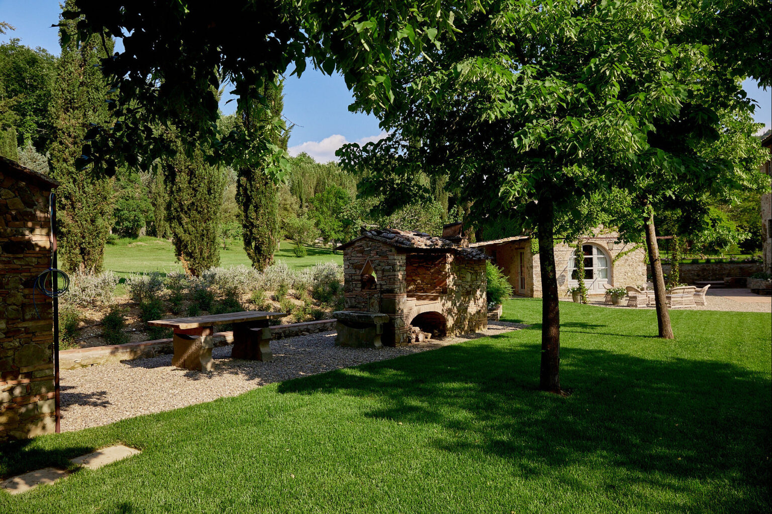 Charming outdoor garden featuring a rustic stone barbecue, wooden bench, and lush greenery, surrounded by tall trees and flowering plants, creating a serene and inviting atmosphere. | Only on SopranoVillas