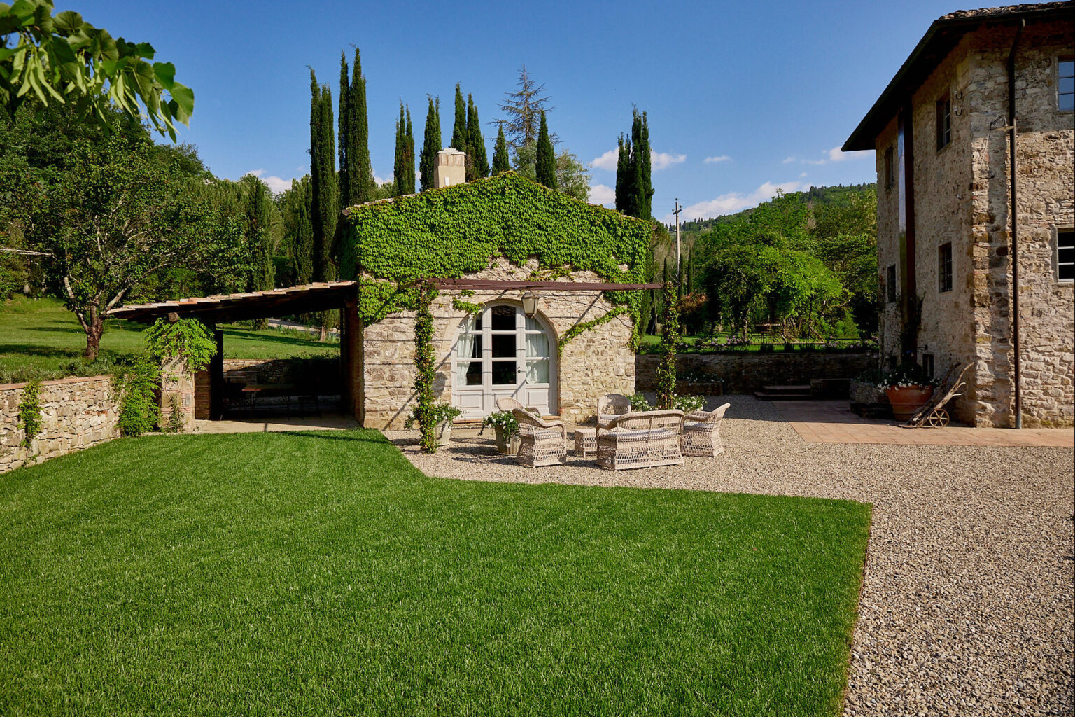 Charming rustic stone cottage surrounded by lush greenery and cypress trees, featuring a cozy outdoor seating area on a gravel patio, perfect for relaxation in a serene landscape. | Only on SopranoVillas