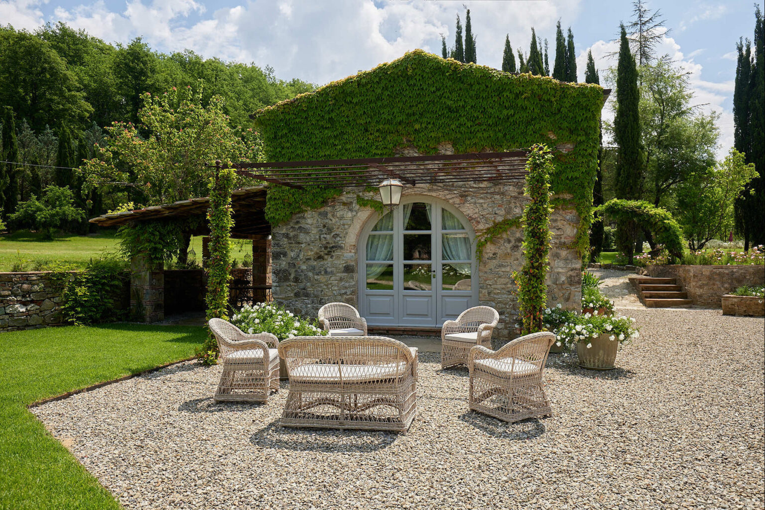 Charming stone cottage surrounded by greenery, featuring a cozy outdoor seating area with wicker chairs on gravel, lush vines climbing the walls, and a serene garden setting. Ideal for relaxation and outdoor gatherings. | Only on SopranoVillas