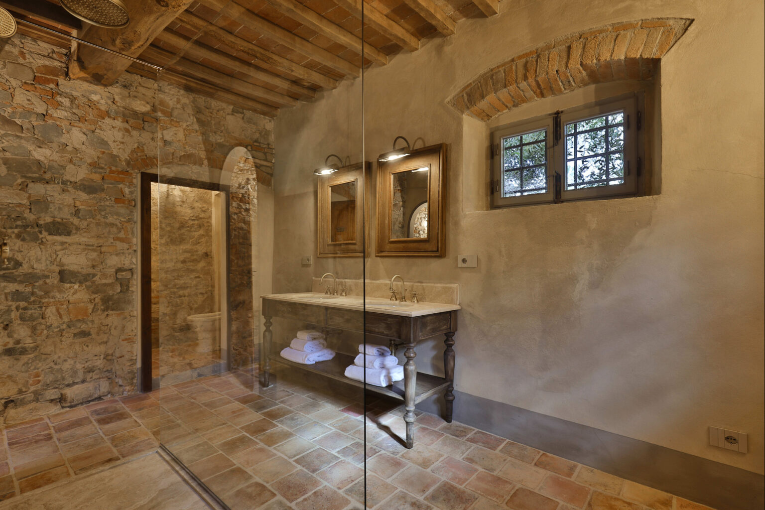 Rustic bathroom interior featuring exposed stone walls, wooden beams, and a glass shower. The space includes a stone vanity with mirrors and neatly arranged towels, creating a warm and inviting atmosphere. | Only on SopranoVillas