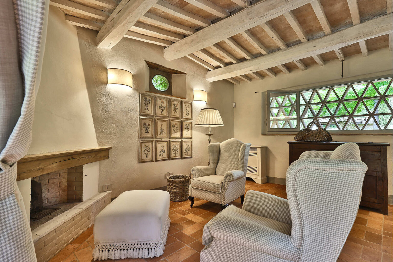 Cozy living room featuring a rustic design with exposed wooden beams, comfortable armchairs, a decorative fireplace, and a unique geometric window. The warm color palette and natural light create an inviting atmosphere, perfect for relaxation. | Only on SopranoVillas