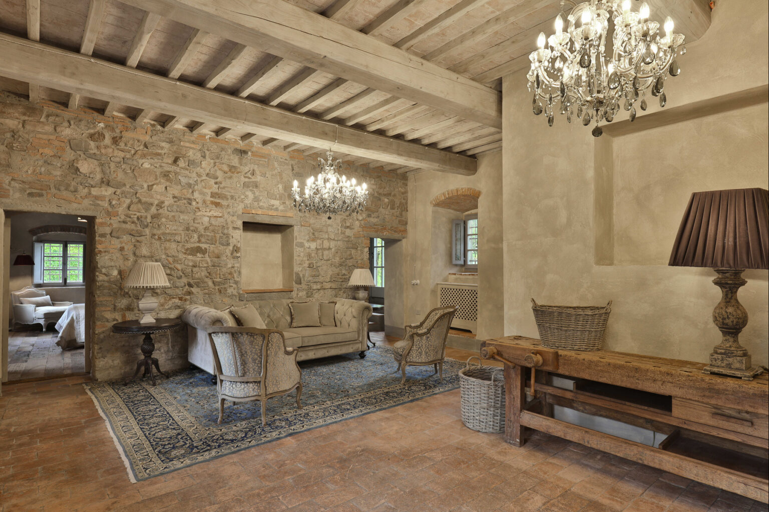 Rustic living room interior featuring exposed stone walls, wooden beams, and elegant chandeliers. The space includes a comfortable sofa, stylish armchairs, and a wooden console table, complemented by a decorative area rug. Natural light filters through the windows, enhancing the cozy ambiance. | Only on SopranoVillas