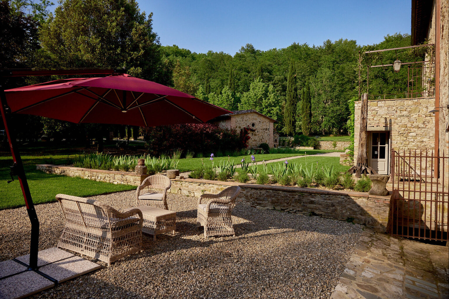 Charming outdoor seating area with wicker chairs and a large red umbrella, surrounded by lush greenery and a landscaped garden featuring stone walls and blooming flowers, ideal for relaxation and enjoying nature. | Only on SopranoVillas