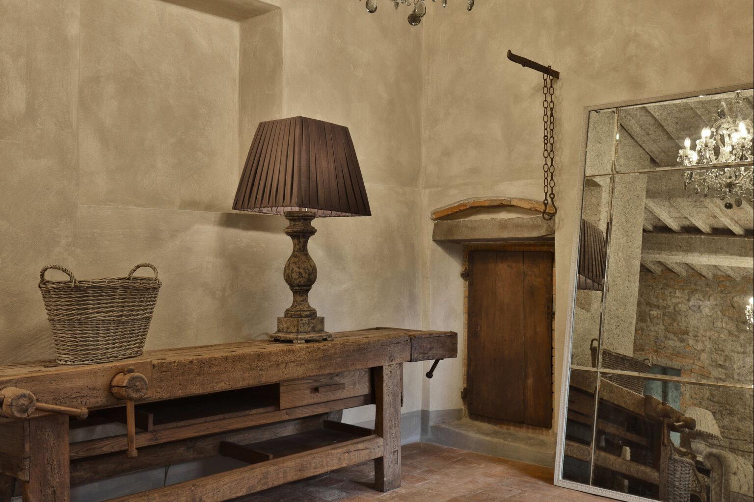 Rustic interior featuring a wooden table with a vintage lamp, a woven basket, and a large mirror. The textured gray walls and warm lighting create a cozy atmosphere, ideal for home decor inspiration. | Only on SopranoVillas
