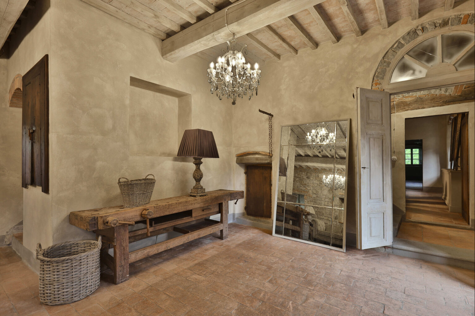 Interior view of a rustic hallway featuring a wooden table, a stylish lamp, a vintage chandelier, and a large mirror, with arched windows and exposed beam ceiling, creating a warm and inviting atmosphere. | Only on SopranoVillas