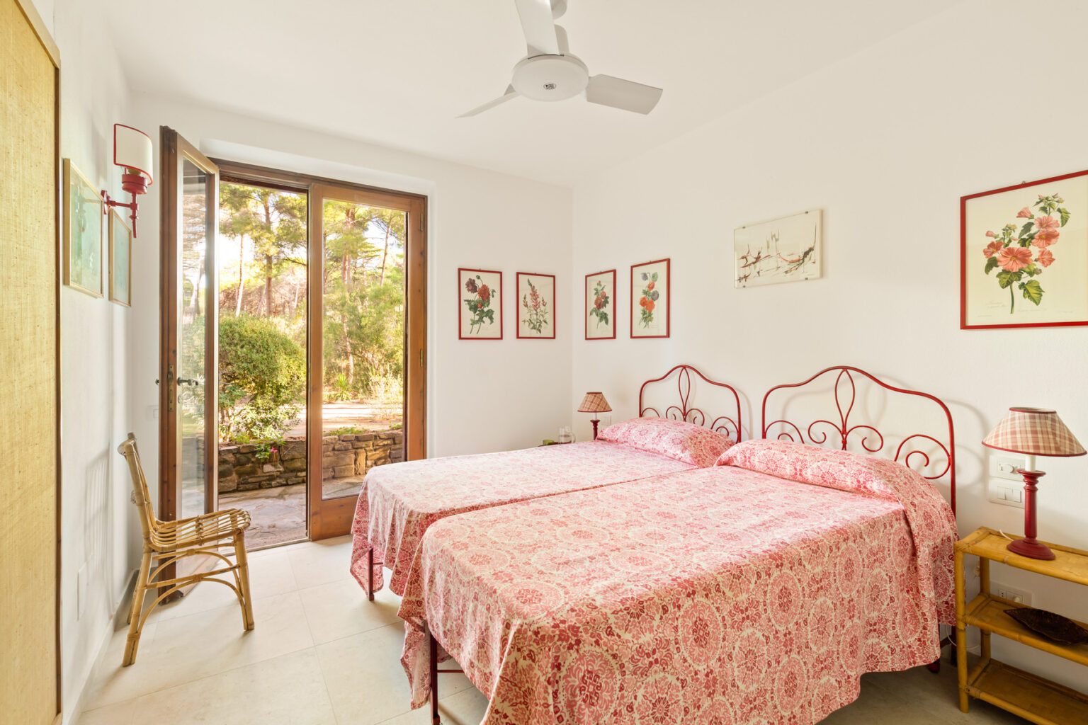 Bright and airy bedroom featuring two twin beds with floral-patterned linens, a ceiling fan, and large windows that open to a garden view, complemented by decorative artwork on the walls. | Only on SopranoVillas