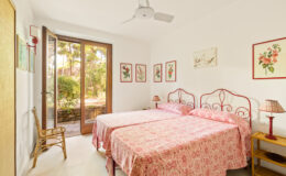Bright and airy bedroom featuring two twin beds with floral-patterned linens, a ceiling fan, and large windows that open to a garden view, complemented by decorative artwork on the walls.