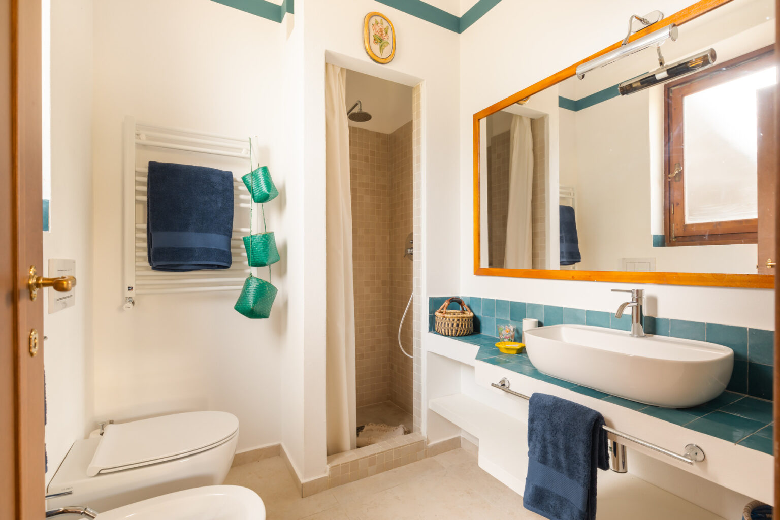 Modern bathroom featuring a walk-in shower, a stylish oval sink, and a large mirror. The space includes a towel warmer with blue towels, green hanging storage baskets, and a wooden window, creating a bright and inviting atmosphere. | Only on SopranoVillas