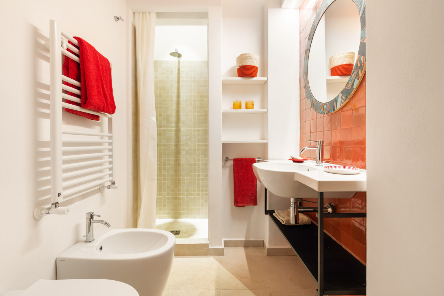 Modern bathroom interior featuring a sleek design with a shower, bidet, and double sink. The space is accented with red towels and decorative items, complemented by a round mirror and warm lighting for a cozy atmosphere. | Only on SopranoVillas
