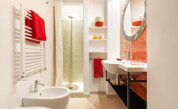 Modern bathroom interior featuring a sleek design with a shower, bidet, and double sink. The space is accented with red towels and decorative items, complemented by a round mirror and warm lighting for a cozy atmosphere.