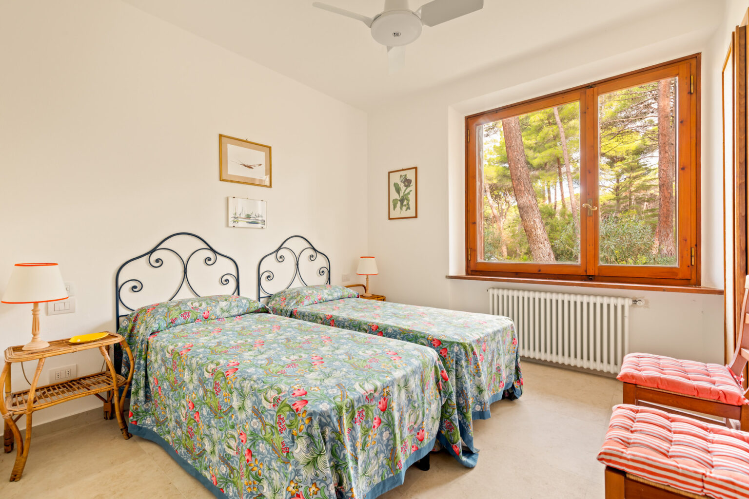 Bright and airy bedroom featuring two twin beds with floral bedspreads, large windows offering a view of greenery, and wooden accents. Ideal for a cozy retreat or guest accommodation. | Only on SopranoVillas