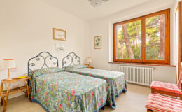 Bright and airy bedroom featuring two twin beds with floral bedspreads, large windows offering a view of greenery, and wooden accents. Ideal for a cozy retreat or guest accommodation.