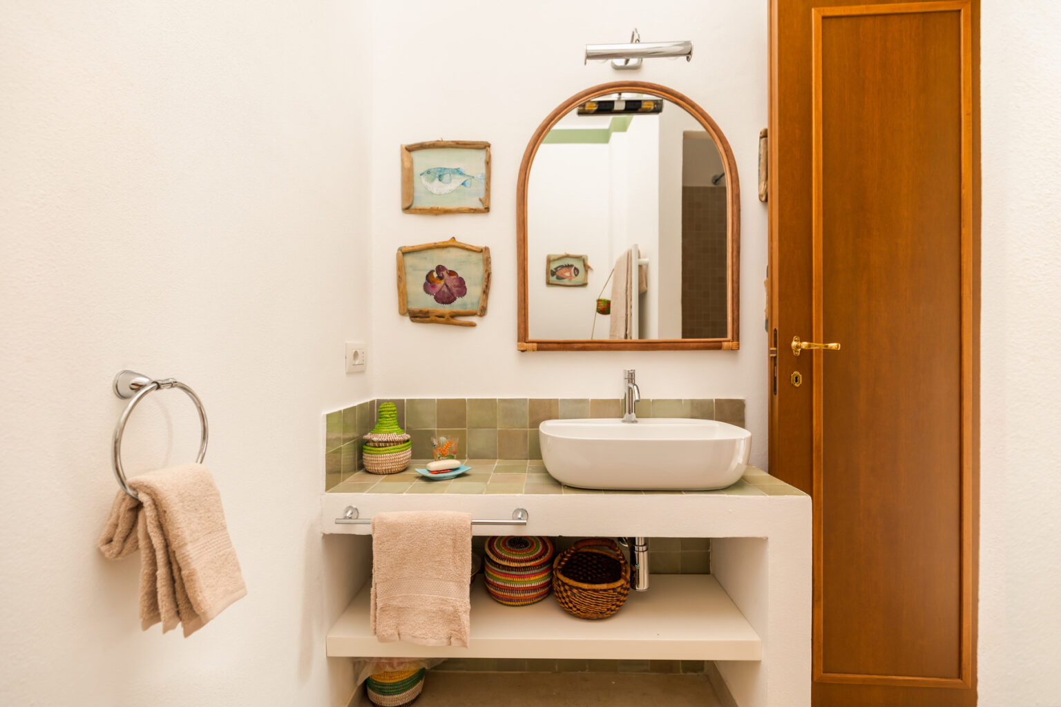 Modern bathroom interior featuring a round mirror above a stylish sink, decorative wall art, and neatly arranged towels. The design includes wooden elements and a clean, minimalist aesthetic. | Only on SopranoVillas