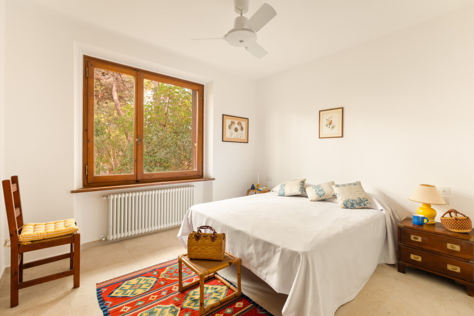 Bright and airy bedroom featuring a double bed with decorative pillows, a wooden chair with a yellow cushion, and a bedside table with a lamp. The room has a large window overlooking greenery, enhancing natural light, and is adorned with framed botanical artwork. A colorful rug adds warmth to the space. | Only on SopranoVillas