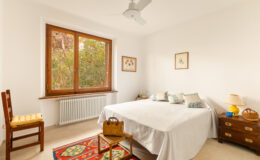 Bright and airy bedroom featuring a double bed with decorative pillows, a wooden chair with a yellow cushion, and a bedside table with a lamp. The room has a large window overlooking greenery, enhancing natural light, and is adorned with framed botanical artwork. A colorful rug adds warmth to the space.