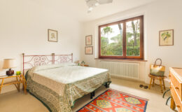 Cozy bedroom featuring a double bed with a patterned bedspread, large window with views of greenery, light-colored walls, wooden furniture, and a decorative rug. Ideal for relaxation and comfort.