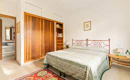 Cozy bedroom featuring a wrought iron bed with a green patterned bedspread, wooden wardrobe, and a side table. The room has a warm, inviting atmosphere with natural light illuminating the space.