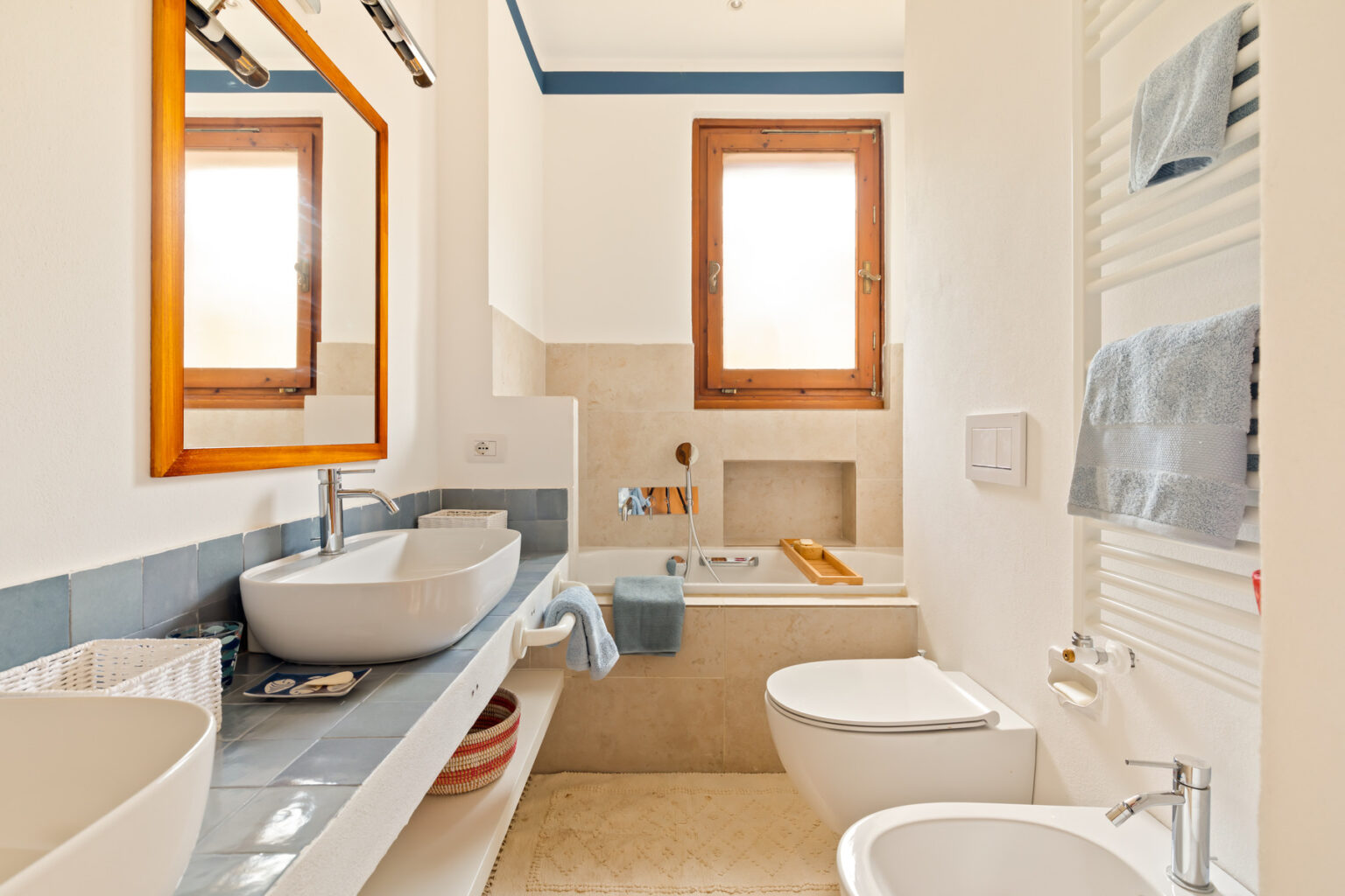 Modern bathroom with natural light, featuring a stylish sink, a bathtub, and a wooden-framed mirror. The space is decorated with blue and neutral tones, creating a calming atmosphere. | Only on SopranoVillas