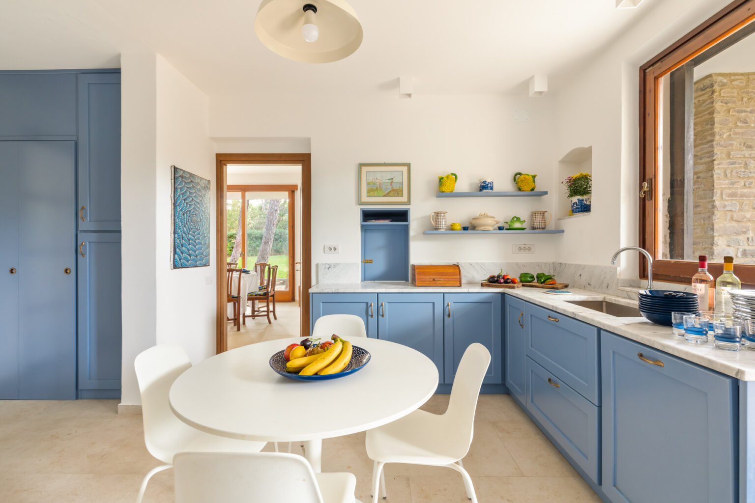 Bright and airy kitchen featuring blue cabinetry, a marble countertop, and a round dining table with a fruit bowl. Shelves display vibrant dishware, and large windows provide natural light and a view of the garden. Modern design elements create a welcoming atmosphere. | Only on SopranoVillas