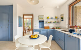 Bright and airy kitchen featuring blue cabinetry, a marble countertop, and a round dining table with a fruit bowl. Shelves display vibrant dishware, and large windows provide natural light and a view of the garden. Modern design elements create a welcoming atmosphere.