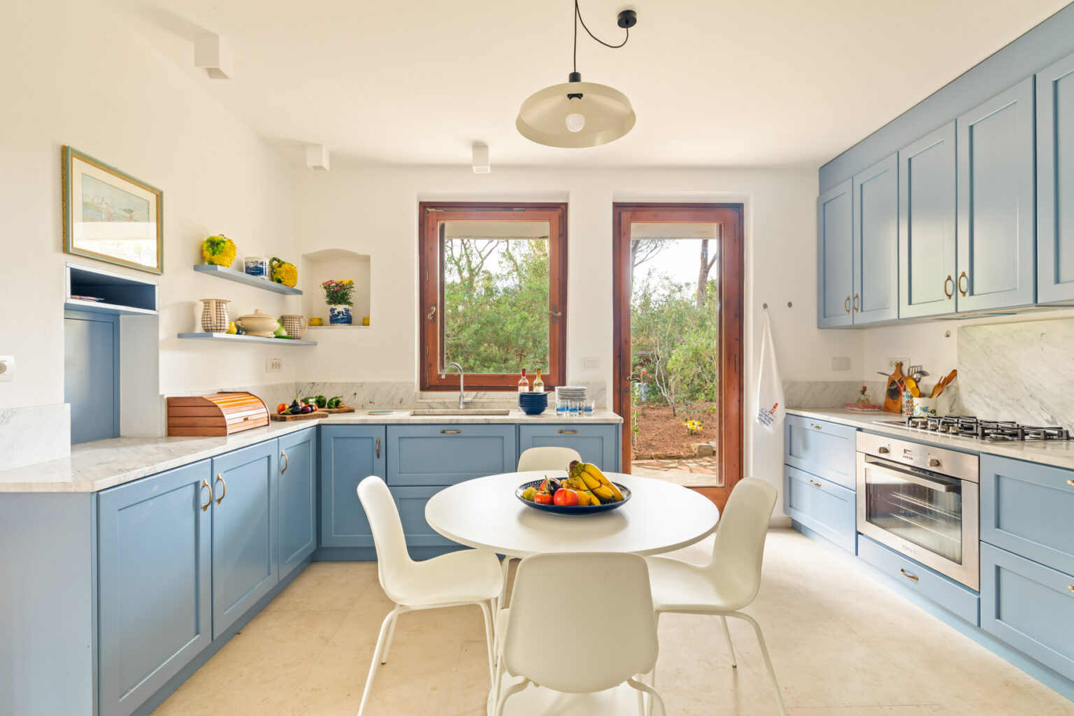 Bright and modern kitchen featuring blue cabinetry, a round dining table with a fruit centerpiece, and large windows that invite natural light. | Only on SopranoVillas