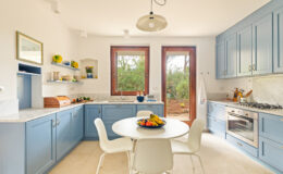 Bright and modern kitchen featuring blue cabinetry, a round dining table with a fruit centerpiece, and large windows that invite natural light.