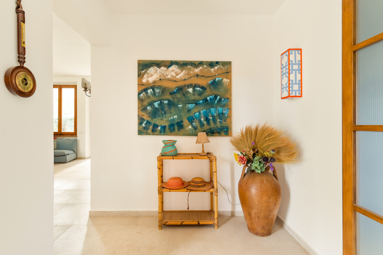 Interior view of a bright entryway featuring a colorful wall art piece, a wooden console table with decorative items, and a large ceramic vase with dried plants, creating a welcoming atmosphere. Natural light floods the space through large windows. | Only on SopranoVillas