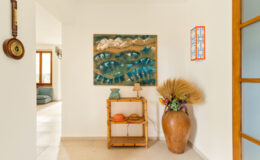Interior view of a bright entryway featuring a colorful wall art piece, a wooden console table with decorative items, and a large ceramic vase with dried plants, creating a welcoming atmosphere. Natural light floods the space through large windows.