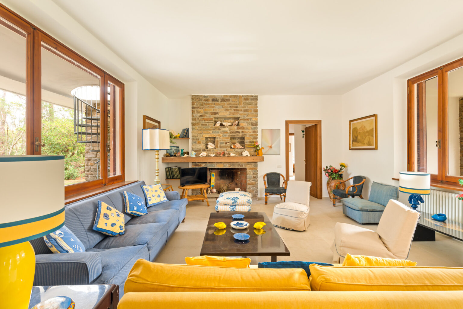 Bright and airy living room featuring a stone fireplace, comfortable blue sofas, and yellow accent pillows. Large windows allow natural light to fill the space, complemented by stylish decor and a cozy atmosphere. | Only on SopranoVillas