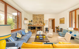 Bright and airy living room featuring a stone fireplace, comfortable blue sofas, and yellow accent pillows. Large windows allow natural light to fill the space, complemented by stylish decor and a cozy atmosphere.