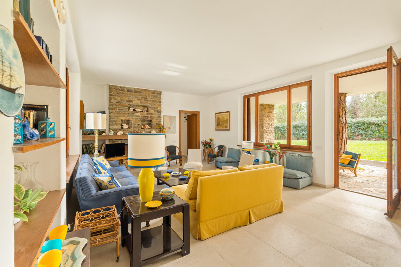 Bright and inviting living room featuring a yellow sofa, blue armchairs, and a stone fireplace. Natural light floods the space through large windows, highlighting decorative elements like plants, artwork, and colorful tableware. A seamless connection to the outdoor area enhances the open and airy atmosphere. | Only on SopranoVillas
