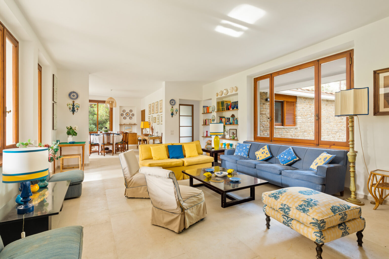 Bright and airy living room featuring a mix of modern and vintage decor. The space includes a yellow sofa, a blue couch with decorative pillows, and a patterned ottoman. Large windows allow natural light, illuminating the light-colored flooring and stylish furnishings. A dining area is visible in the background, showcasing a wooden table and chairs, complemented by wall art and shelves filled with books. | Only on SopranoVillas
