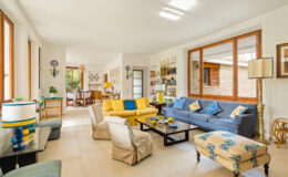 Bright and airy living room featuring a mix of modern and vintage decor. The space includes a yellow sofa, a blue couch with decorative pillows, and a patterned ottoman. Large windows allow natural light, illuminating the light-colored flooring and stylish furnishings. A dining area is visible in the background, showcasing a wooden table and chairs, complemented by wall art and shelves filled with books.
