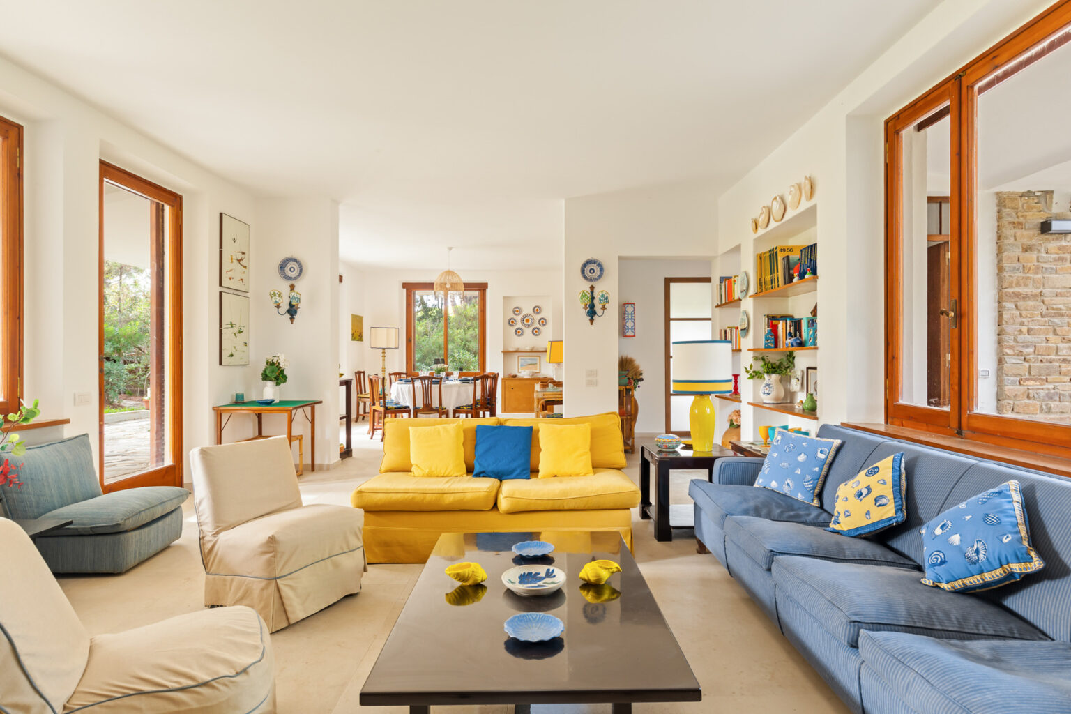 Bright and airy living room featuring a mix of yellow and blue sofas, decorative pillows, and large windows allowing natural light. The space includes a coffee table with decorative items, a cozy armchair, and a dining area visible in the background. Ideal for modern home decor inspiration. | Only on SopranoVillas