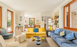 Bright and airy living room featuring a mix of yellow and blue sofas, decorative pillows, and large windows allowing natural light. The space includes a coffee table with decorative items, a cozy armchair, and a dining area visible in the background. Ideal for modern home decor inspiration.