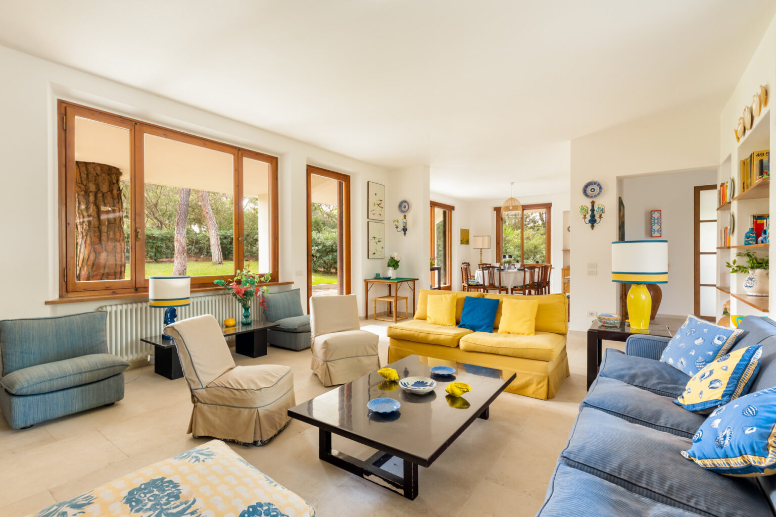 Bright and spacious living room featuring a mix of comfortable seating, including a blue sofa and beige armchairs, a modern coffee table, and large windows that let in natural light. The decor includes vibrant yellow accents and plants, creating a welcoming atmosphere. | Only on SopranoVillas