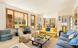 Bright and spacious living room featuring a mix of comfortable seating, including a blue sofa and beige armchairs, a modern coffee table, and large windows that let in natural light. The decor includes vibrant yellow accents and plants, creating a welcoming atmosphere.
